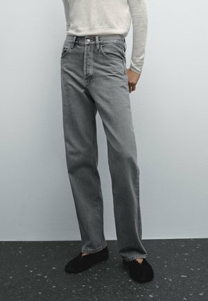 Persoon draagt grijze jeans met hoge taille en rechte pijpen, witte top met lange mouwen en zwarte instapschoenen, staand tegen een lichtgrijze muur.