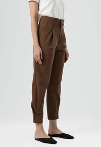 Bruine broek met hoge taille en taps toelopende pasvorm, voorzien van plooien aan de voorkant en een knoopdetail aan de manchet, gecombineerd met zwarte instapschoenen.