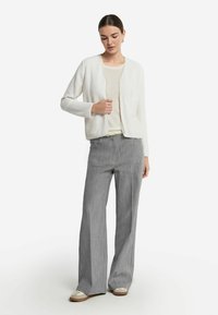 Weißer strukturierter Cardigan mit Rundhalsausschnitt, kombiniert mit hellgrauen weitbeinigen Hosen. Die Schuhe zeigen eine Farbgestaltung in Creme und Braun.