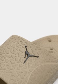 Μπεζ παντόφλα με υφασμάτινη επιφάνεια, με λογότυπο Jumpman σε μαύρο χρώμα. Κατασκευασμένη από ευέλικτο υλικό, σχεδιασμένη για άνεση και ευκολία.