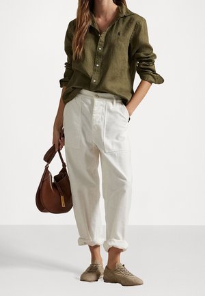 Vrouw draagt olijfgroen shirt met lange mouwen, witte opgerolde broek, beige sneakers, houdt bruine handtas vast met één hand in de zak.
