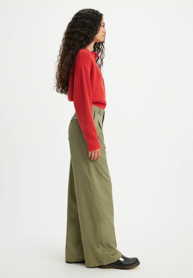 Pull rouge en tricot, pantalon vert à jambe large et sabots noirs. Texture côtelée prononcée sur le pull et taille ajustée sur le pantalon.