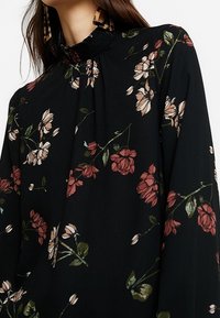 Svart blommig blus med långa ärmar, med ett rynkat halsen och spridda rosa och vita blommönster på en mörk bakgrund.