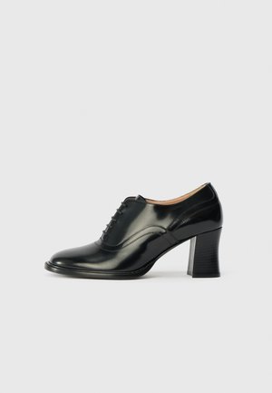 Des chaussures à talons aiguilles en cuir noir avec un bout arrondi, un design à lacets et des coutures subtiles, ainsi qu'un talon empilé texturé.
