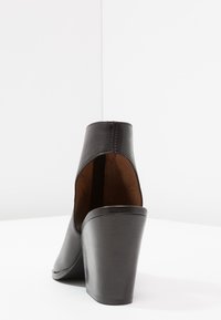 Czarna skórzana szpilka z zamkniętym tyłem i otwartymi bokami, widoczna z tyłu na białej powierzchni i tle.