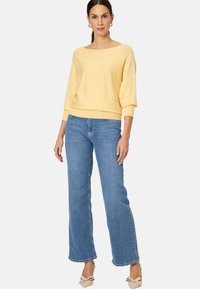 Gelber, lässig geschnittener Pullover mit weitem Ausschnitt und hellblauen, weiten Jeans; hat eine strukturierte Oberfläche und dezente Saumdetails.