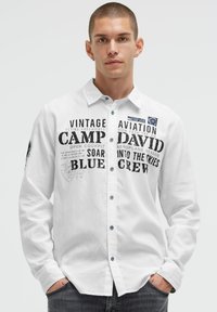 Weißes Langarmhemd mit fettem schwarzen Grafiktext und Marineblau-Akzenten, mit Button-down-Kragen und blauen Knöpfen auf der Vorderseite.