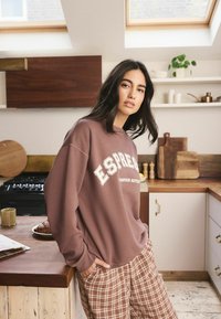 Brauner Sweatshirt mit weißem Text, lässiger Schnitt. Kombiniert mit karierten, weit geschnittenen Schlafanzughosen. Küchensetting mit Holzakzenten.