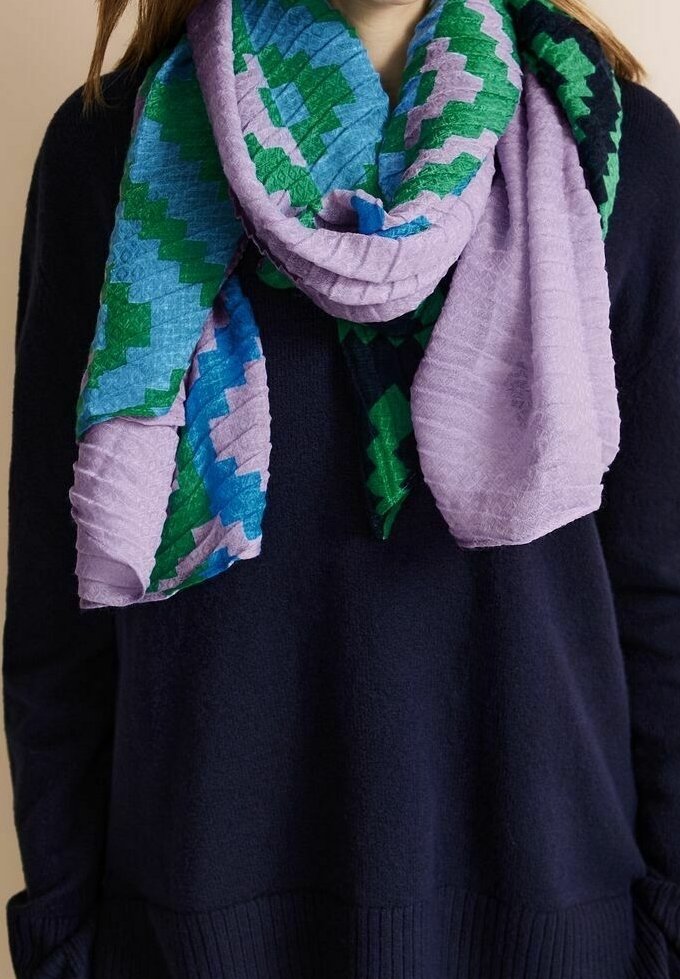Personne portant un pull noir avec une écharpe texturée aux motifs en zigzag verts, bleus, violets et lavande enroulée autour du cou.