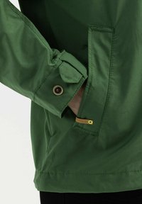 Manche de veste verte avec poignet boutonné et main partiellement dans la poche latérale avec une petite étiquette marron indiquant "Camel Active".