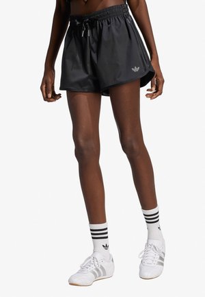 Persona snella che indossa pantaloncini neri Adidas, calzini bianchi Adidas con strisce nere e scarpe da ginnastica bianche, in piedi davanti a uno sfondo bianco.