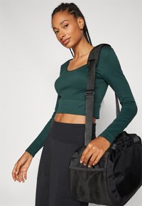 Grønn ribbet langermet crop-top kombinert med mørke bukser. Modellen bærer en svart treningsbag med en teksturert nettinglomme.