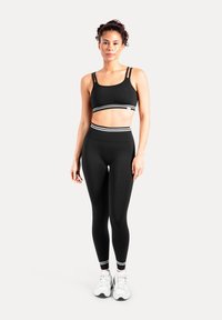 Svart sport-bh med tunna band och ett vitt randigt band, kombinerad med svarta högmidjade leggings med matchande muddar. Vita sneakers.