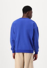 Felpa di cotone blu con vestibilità rilassata, polsini a costine e collo a giro. Texture liscia; indossata con pantaloni beige chiaro.