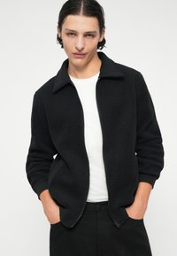 Giacca in pile nera con zip frontale, maniche lunghe e colletto. Il modello indossa una maglietta bianca sotto. La texture è morbida e soffice.