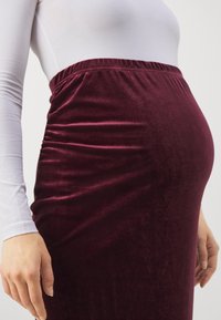 Burgundy sammet graviditetskjol med elastisk midja, med en slät textur och rynkade detaljer längs sidorna, designad för komfort.