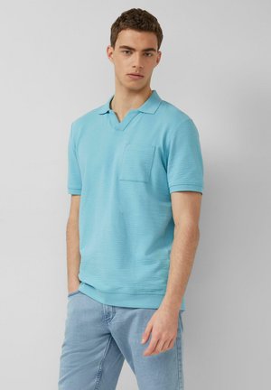 Młody mężczyzna o krótkich brązowych włosach, ubrany w jasno niebieską teksturowaną koszulkę polo i jasne niebieskie dżinsy, stojący na tle jednolitej szarej ściany.