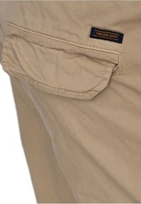 Pantalons en toile beige avec une poche arrière à rabat. La poche comporte une étiquette bleu marine avec "Indicode Jeans" en texte orange.