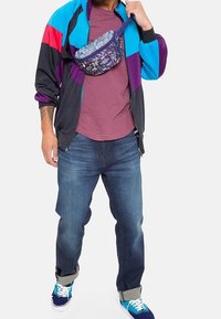 Man som bär en färgglad träningsjacka, mörkröd t-shirt, blå jeans, blå sneakers och en blommig midjeväska över bröstet.