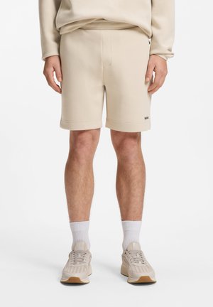 Mann trägt beige legere Shorts, kombiniert mit passenden Sneakers und weißen Sportsocken, steht vor einem einfarbigen Hintergrund.