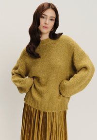 Senfgelber Strickpullover mit überschnittenen Schultern, geripptem Saum und weicher Textur, kombiniert mit einem golden plissierten Rock.