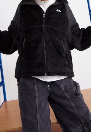 Persona con chaqueta polar negra con cremallera y pantalones oscuros y sueltos, de pie con los brazos ligeramente levantados en el interior.