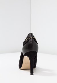 Chaussure à talons hauts noire avec une finition en daim et des accents en cuir lisse. Présente un détail tressé en or près du talon et une pointe effilée.