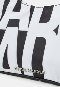 Gros plan d'un sac en tissu blanc et noir à motifs avec un logo métallique argenté "Steve Madden" près du bord inférieur.