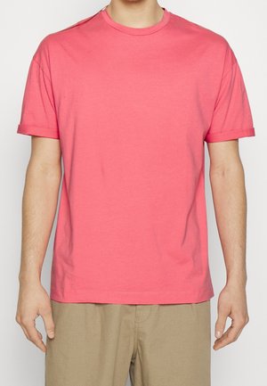 Mand iført en ensfarvet koralrosa T-shirt med korte ærmer og opsmøgede manchetter samt beige bukser, vist fra skuldre til hofter.