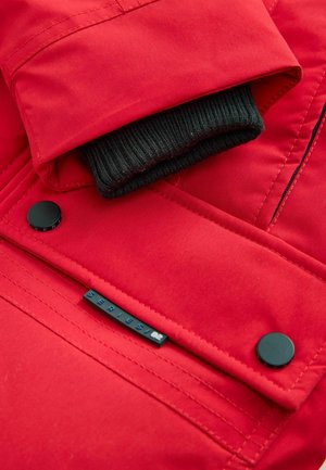 Rood gewatteerd jack met zwarte geribde manchetten. Voorzien van een zwarte drukknoop en een label met "SERIES 82." Gladde, iets glanzende textuur.