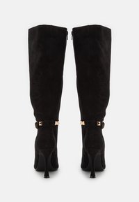 Botas de ante negras hasta la rodilla con punteras puntiagudas, tacones stiletto delgados y hebillas de acento doradas en los tobillos. Textura suave en toda su superficie.