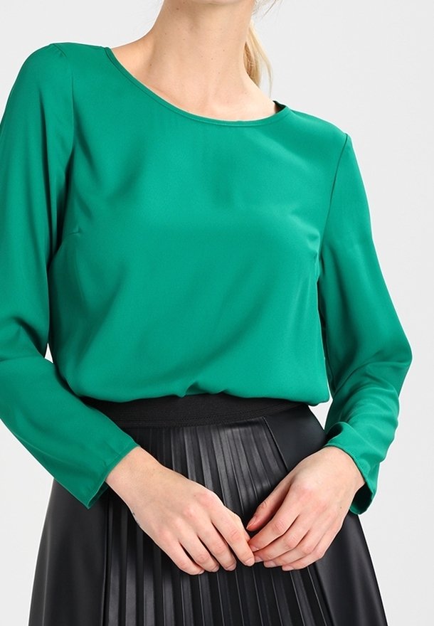 Femme portant un chemisier vert à manches longues rentré dans une jupe noire plissée taille haute, les mains délicatement jointes à la taille.