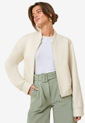 Giacca in pile color crema con chiusura a zip, polsini a costine e colletto, abbinata a pantaloni verdi a vita alta.