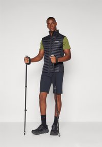 Black padded vest, green short-sleeve shirt, dark shorts, and black hiking shoes with trekking poles. Smooth texture and fitted design.