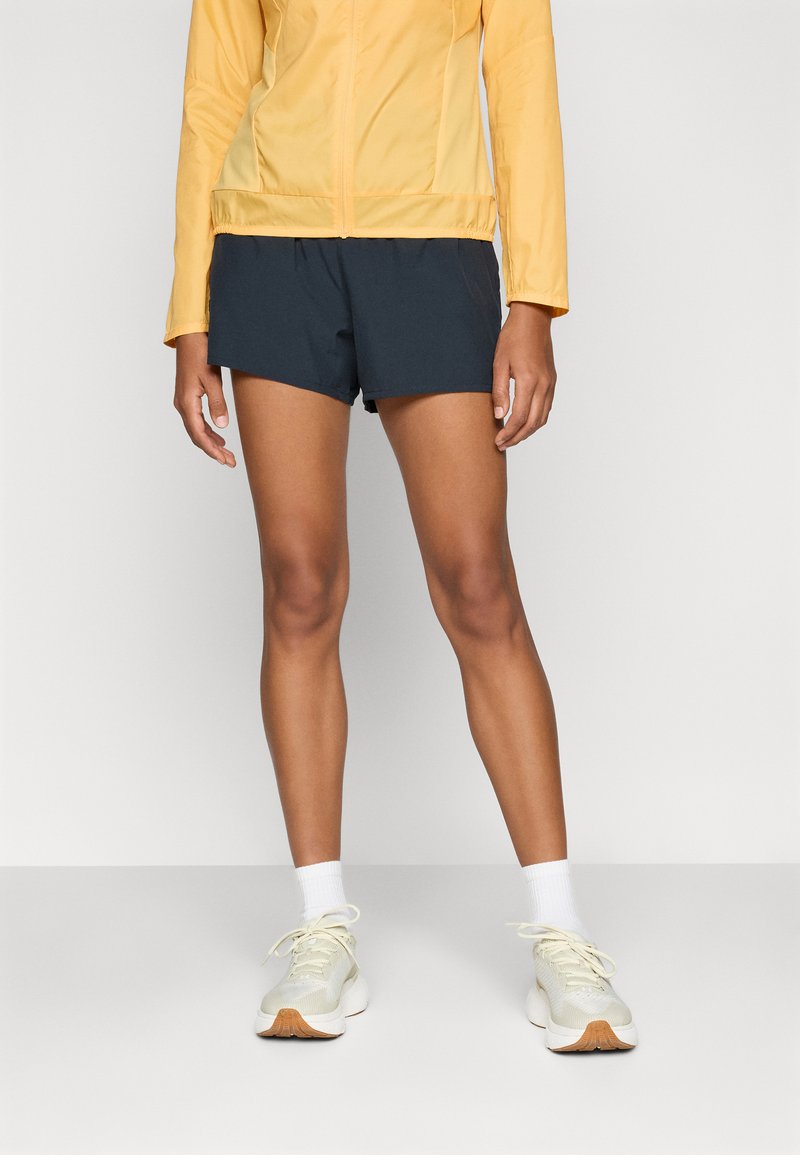 Veste légère jaune à manches longues, shorts marins, et chaussures de sport crème avec une finition texturée et des semelles de couleur claire.