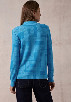 Suéter azul con un patrón geométrico texturizado. Mangas largas, diseño con cuello y puños acanalados. Combinado con jeans oscuros.