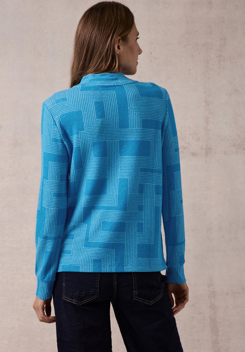 Blauer Pullover mit einem strukturierten geometrischen Muster. Lange Ärmel, modern geschnittenes Design mit Kragen und gerippten Bündchen. Kombiniert mit dunklen Jeans.