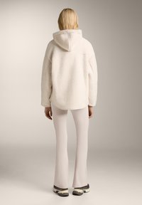 Felpa con cappuccio in beige chiaro, con una vestibilità rilassata, abbinata a pantaloni a zampa di elefante rosa chiaro e sneakers sportive. Texture morbida.