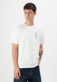 White cotton t-shirt with short sleeves, crew neck, featuring a small blue Timberland logo on the left side. Worn with dark blue jeans.