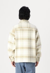 Fleece skjortejakke i creme med beige og grå tartanmønster. Har lange ærmer, krave og en afslappet pasform. Tekstureret stof.
