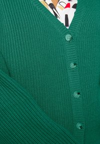 Grön stickad cardigan med V-hals, sex matchande knappar, ribbad textur och ett synligt mönstrat foder.