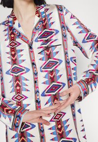 Chemise en soie ornée d'un motif tribal vibrant en rouge, bleu et noir, avec des poignets contrastés et un devant boutonné. Comprend une poche sur la poitrine.