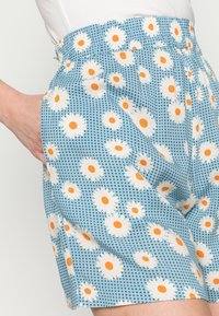 Siniset shortsit, joissa on ruudukkomalli, valkoisia ruiskaunokkeja oransseilla keskuksilla. Kangas on kevyt, ja shortsien vyötäröllä on joustava nauha sekä sivutaskut.