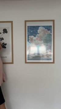 Rėmelis su plakatu, kuriame pavaizduoti spalvingi debesys, mėlyna dangus ir tolumoje matoma bokšto užuomazga. Medinis rėmas; tekstas pateiktas keliose kalbose.