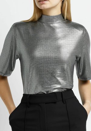 Frau trägt ein glänzendes silbernes Kurzarm-Oberteil mit Stehkragen, das in hoch taillierte schwarze Hosen gesteckt ist, mit den Händen in den Taschen.