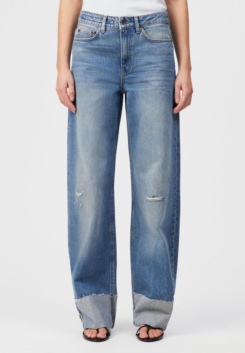 Lyseblå vide jeans med slitte detaljer nær knærne og frynsete vide mansjetter, kombinert med svarte sandaler med åpen tå og hvit topp.