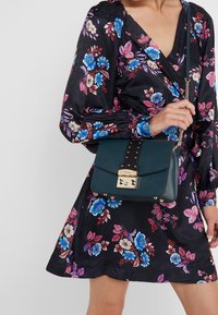 Donna che indossa un vestito floreale nero mentre tiene una borsa verde scuro con chiusura e tracolla dorata attraversata sul corpo.