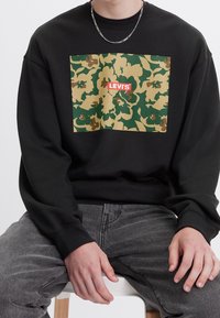Felpa nera con stampa camouflage a rettangolo che presenta il logo "LEVI'S" in rosso. Materiale in misto cotone, polsini a costine e vestibilità casual.