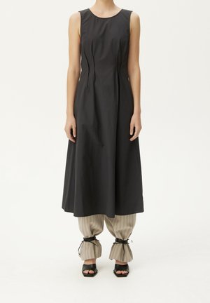 Robe midi noire sans manches portée sur un pantalon beige à fines rayures, rassemblé et noué aux chevilles, associée à des sandales noires à bout ouvert.