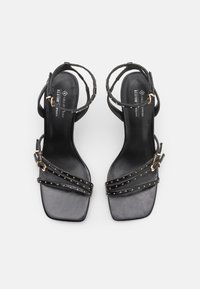 Black vegan leather high-heeled sandals featuring multiple thin straps with gold studs, a square toe, and adjustable ankle buckle.